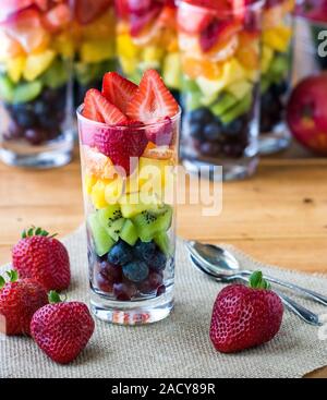 Una vista ravvicinata di un arcobaleno parfait di frutta in un bicchiere alto circondato da fragole e diversi rainbow parfaits in background. Foto Stock