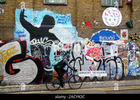 Un ciclista passando graffiti su un muro di mattoni in Buxton Street off Brick Lane Whitechapel Londra Inghilterra REGNO UNITO Foto Stock