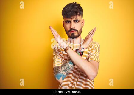 Giovane uomo con tatuaggio a strisce che indossa la t-shirt in piedi isolato su sfondo giallo rifiuto espressione bracci di incrocio facendo segno negativo, arrabbiato f Foto Stock