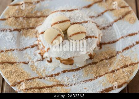 Splendida mattina, ottima colazione.deliziose frittelle di banane e gelato alla vaniglia, sciroppo dolce, amaretti , scaglie di cocco sulla piastra bianca e rustico in legno tabella.Top view.laici piana. Foto Stock