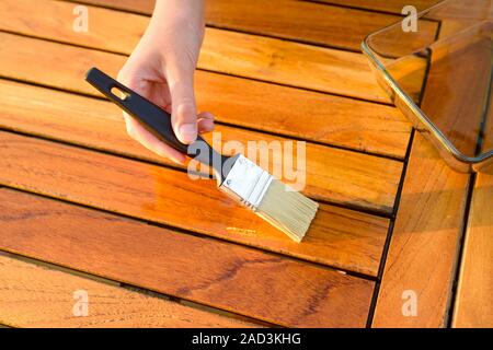 Mano che tiene un pennello applicare vernice vernice su una tavola di legno Tavolo da giardino - pittura e manutenzione legno olio-cera Foto Stock