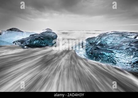 Blocchi di ghiaccio in sfumature di blu su una spiaggia nera con forte surf, il movimento di acqua può essere visto (lunga esposizione, cancellare le tracce di movimento) Foto Stock