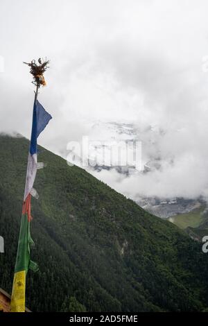 Un snow-capped picco in Himalaya picchi attraverso le nuvole mentre le bandiere di preghiera vola nel vento. Il canto di un uccello e il suo nido resto in cima il flag. Foto Stock