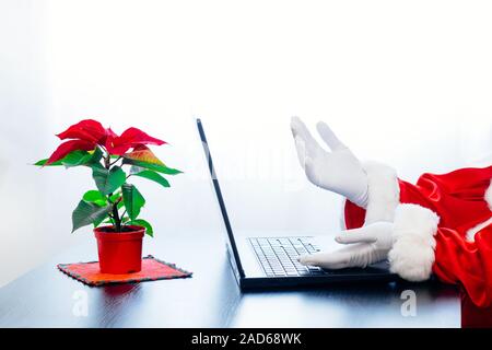 Babbo Natale utilizzando laptop su sfondo bianco con red poinsettia. Vestito in guanti bianchi mani di Santa la digitazione sul computer. Foto Stock