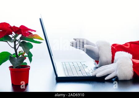 Babbo Natale utilizzando laptop su sfondo bianco con red poinsettia. Vestito in guanti bianchi mani di Santa la digitazione sul computer. Foto Stock