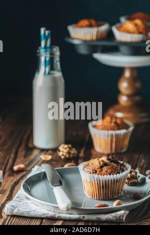 Vaniglia e caramello muffin in bicchieri di carta e bottiglie di latte sul legno scuro dello sfondo. Deliziosa tortina con uvetta, mandorle e noci. In casa biscui Foto Stock