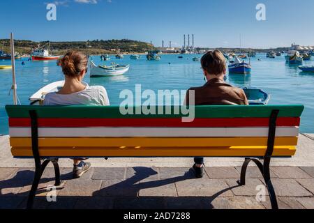 Due visitatori di Marsaxlokk villaggio di pescatori seduti su un dipinto luminosamente panca su il porto la luce del sole. Malta, Mediterranea. Foto Stock