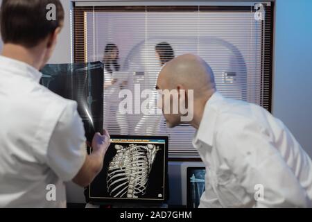 Nella sala di controllo medico radiologo e discutere della diagnosi mentre si guarda la procedura, in Background subisce paziente MRI o CT Procedura di scansione. Foto Stock