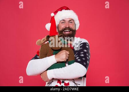 Giocattolo morbido. Abbraccio e l'amore. Acquistare regali. Inverno vendita. Festa d'inverno. La renna. L'uomo santa hat tenere regalo di Natale sfondo rosso. Natale concetto prep. Tanga faccia barbuta inverno maglione. Vacanze invernali. Foto Stock