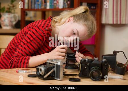 Ragazza adolescente servendosi di un piccolo cacciavite per riparare un antico fotocamera a pellicola. Altre telecamere sono porre sul tavolo di fronte a lei. Foto Stock