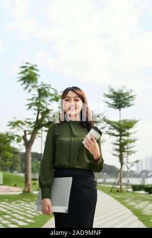 Piuttosto giovane donna che porta una tazza di caffè da asporto e un file in un parco urbano come ella offre alla telecamera un sorriso smagliante Foto Stock