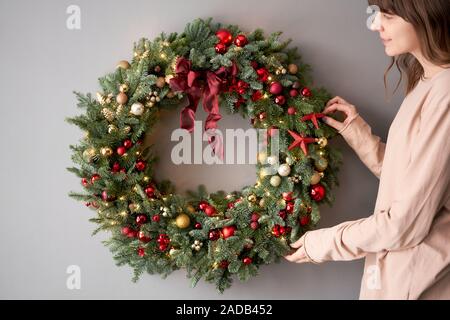 Bella festosa ghirlanda di abete rosso fresco nelle mani della donna. Xmas circlet con rosso e oro, ornamenti e sfere. Il Natale di umore. Muro grigio sullo sfondo. Foto Stock