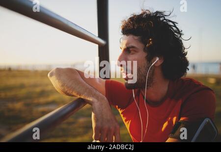 Vista laterale di un esaurito giovane atleta maschio ascoltando musica su auricolari appoggiata su barre in palestra park che guarda lontano verso il tramonto Foto Stock