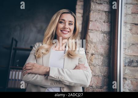 Foto di affascinanti toothy boss raggiante signora con mani giunte indossando blazer sorridendo vicino alla finestra in ufficio della sua azienda Foto Stock