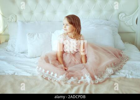 Poco bella ragazza in un abito rosa siede su un letto in camera da letto e guarda fuori dalla finestra. Foto Stock