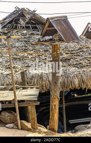 Il vecchio spirito di legno casa con rusty tetto sorge trascurato al sole con un palm-tetto di paglia casa sullo sfondo. Foto Stock