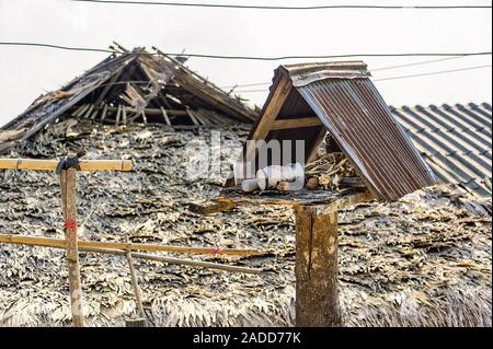 Il vecchio spirito di legno casa con rusty tetto sorge trascurato al sole con un palm-tetto di paglia casa sullo sfondo. Foto Stock