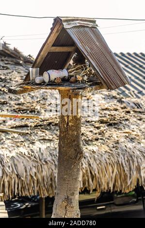 Il vecchio spirito di legno casa con rusty tetto sorge trascurato al sole con un palm-tetto di paglia casa sullo sfondo. Foto Stock