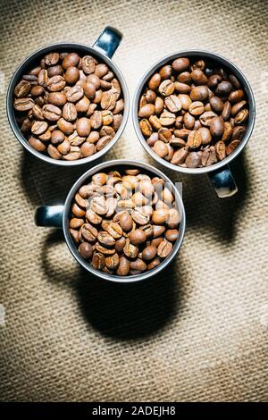 Immagine di overhead di tre tazze di metallo pieno di caffè torrefatto in grani su un sfondo di Hesse. Foto Stock