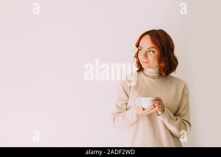 Stile di vita ritratto di dai capelli rossi bella donna sorseggiando il caffè da una tazza bianca, indossa un maglione accogliente. Guardando a parte. Copia dello spazio. Foto Stock