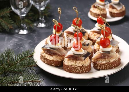 Crostini con le aringhe salate, formaggio, uova di quaglia e pomodori ciliegia su crostoni di pane di segale sullo sfondo di Natale Foto Stock