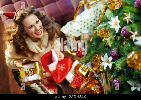 Sorridente donna moderna in oro zecchino gonna e maglione bianco azienda strappata regalo di Natale scatola con crema per il viso che appena uscito da una calza unde Foto Stock