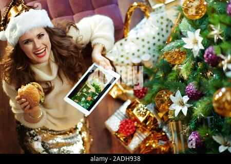 Sorridente donna moderna con lunghi capelli bruna in oro zecchino gonna e maglione bianco sotto albero di Natale decorato nei pressi di caselle presenti a mangiare un hamburger Foto Stock