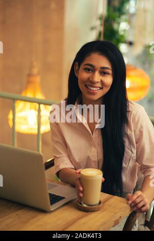 Fiducioso freelancer seduto con il latte nel caffè e sorridente Foto Stock