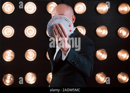 Incentrato sulla fotografia di carte da gioco. Mago in abito nero in piedi in camera con illuminazione speciale al backstage Foto Stock