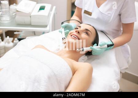 Excited donna giacente in asciugamano bianco durante la terapia microcurrent Foto Stock