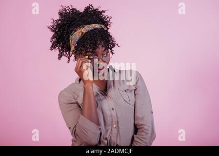 Cercando su nuovi occhiali. Attraente afro american donna in abiti casual a sfondo rosa in studio Foto Stock