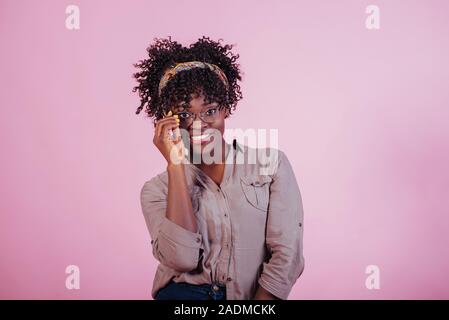 Guardando nella telecamera. Cercando su nuovi occhiali. Attraente afro american donna in abiti casual a sfondo rosa in studio Foto Stock