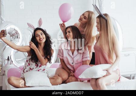 Cattura del momento. Addio al nubilato di partito. Quattro ragazza in rosa e bianche vesti si siede sul divano e prendere selfie Foto Stock