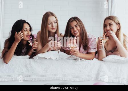 Bere del buon cocktail. Gioiosa le ragazze in calzature sdraiato sul letto in camera bianca e hanno la celebrazione Foto Stock