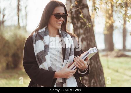 Meteo gioiosa. Giovani sorridente brunette in occhiali da sole sorge nel parco vicino agli alberi e mantiene il blocco note Foto Stock