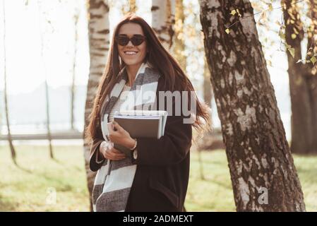 Bel ritratto. Giovani sorridente brunette in occhiali da sole sorge nel parco vicino agli alberi e mantiene il blocco note Foto Stock