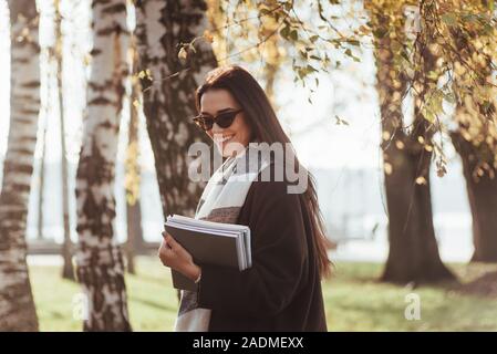 I giorni caldi. Giovani sorridente brunette in occhiali da sole sorge nel parco vicino agli alberi e mantiene il blocco note Foto Stock