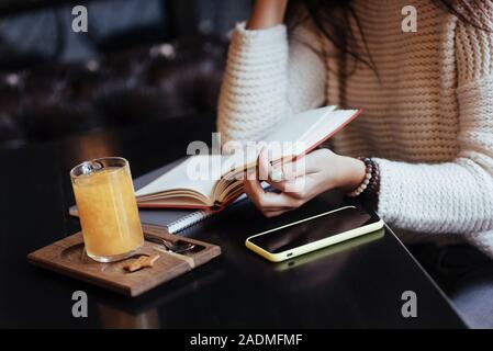 Alcune nuove informazioni. Tagliato vicino la foto della donna leggere il libro nel ristorante con drink di giallo e lo smartphone su tavola Foto Stock