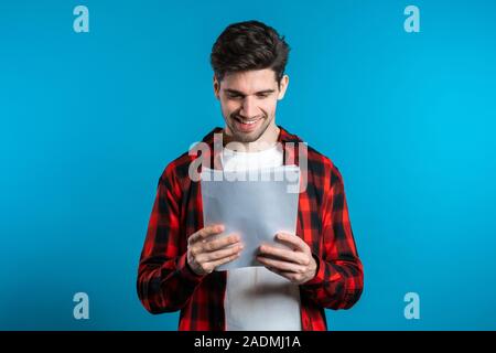 Giovane uomo bello controlla i documenti, bollette, relazione. Imprenditore isolato su sfondo blu. È soddisfatto con il lavoro del personale aziendale. Foto Stock