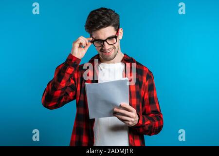Giovane uomo bello controlla i documenti, bollette, relazione. Imprenditore isolato su sfondo blu. È soddisfatto con il lavoro del personale aziendale. Foto Stock