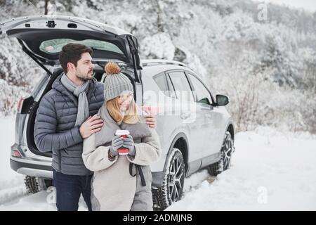 Il freddo ma caldo cuori. Ritratto di giovane coppia che sta insieme vicino alla moderna argento auto presso il nevoso inverno boschi Foto Stock