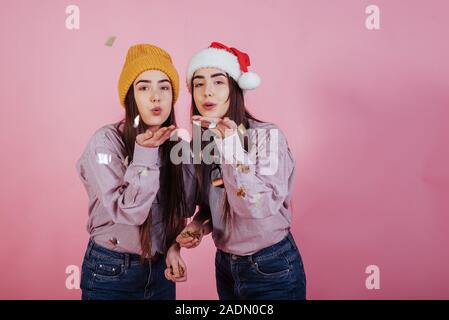 È tempo di Natale. Coriandoli e buon umore per tutti. Baci d'aria. Due sorelle gemelle in piedi e in posa in studio con sfondo rosa Foto Stock