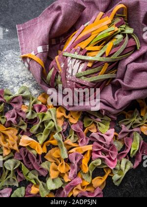 Varietà di pasta cruda, tipi di farfalle bow tie-, fettuccine o tagliatelle pasta colorata su uno sfondo grigio con farina Foto Stock