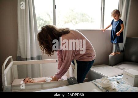 Mom cambiando pannolino di un neonato con big sister permanente sulla sedia Foto Stock