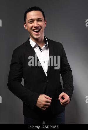 Happy Fun giovane uomo d affari di ridere in abito nero abbigliamento su grigio scuro dello sfondo con copia vuota dello spazio. Closeup ritratto Foto Stock