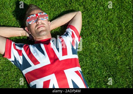 Rilassata giovane britannico in Unione Jack T-shirt e occhiali da sole si trova indietro su un soleggiato patch di estate verde erba Foto Stock