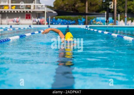 Una donna di nuoto in una piscina pubblica con una nuotata giallo cappello e occhiali facendo il freestyle crawl anteriore corsa nuoto Foto Stock