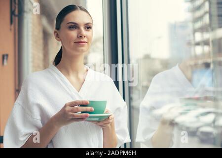 Bella donna godendo il suo tè mentre sta in piedi vicino alla finestra Foto Stock