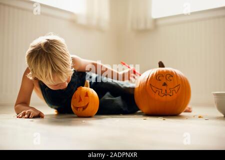 Ragazzo giovane si trova sul suo lato e guarda verso il basso in corrispondenza di una piccola Jack O'Lanterna accanto ad un grande, uncarved zucca con una faccia disegnata su di esso. Foto Stock