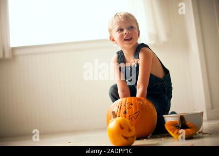 Ragazzo cerca e sorrisi come egli si inginocchia su un pavimento e scava entrambe le mani in una grande zucca con una faccia disegnata su di esso con una ciotola e un piccolo finito Jack O'Lanterna con una candela accesa all'interno davanti a lui. Foto Stock